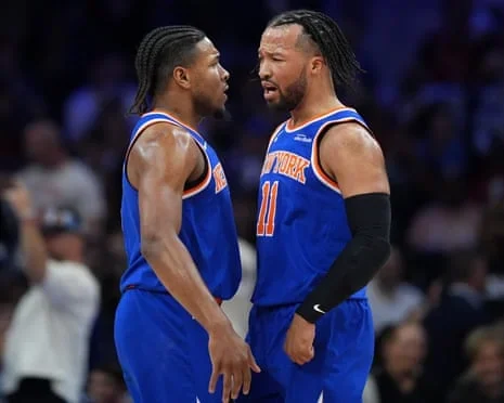 Miles McBride and Jalen Brunson in blue Knicks jerseys face each other in close conversation on court.